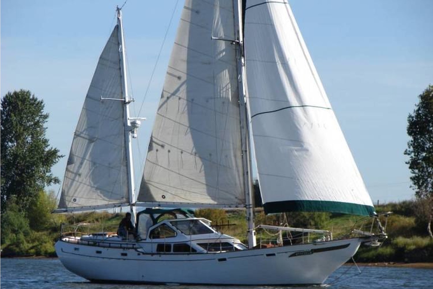 Shawnee, a white Hardin 44 ketch, under full sail on a river lined with trees